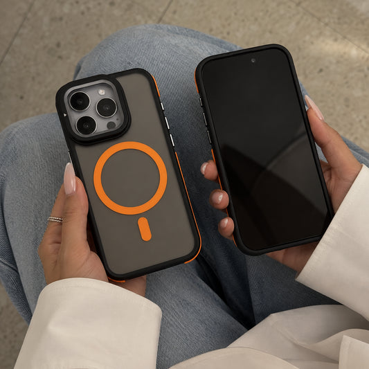 A person holds two smartphones in their hands, one showing a dark gray phone case with a prominent orange MagSafe ring and the other showing a black phone case. The person is wearing blue jeans and a white long-sleeve shirt, with a neutral stone floor in the background.