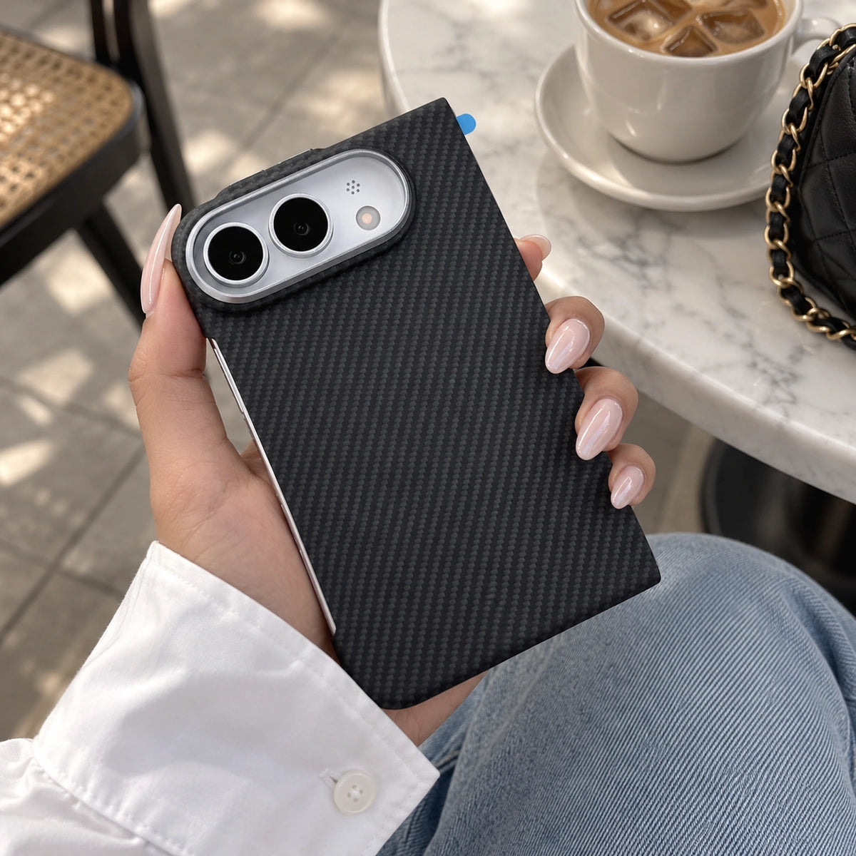 A person holds a smartphone with a black carbon-fiber-textured case in a cafe setting. The case has a silver camera surround and is shown against a background of a white marble table, a coffee cup, and a black quilted bag with a gold chain.