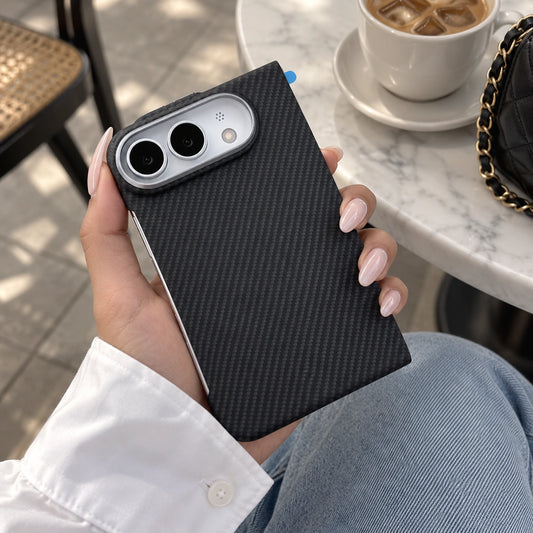 A person holds a smartphone with a black carbon-fiber-textured case in a cafe setting. The case has a silver camera surround and is shown against a background of a white marble table, a coffee cup, and a black quilted bag with a gold chain.