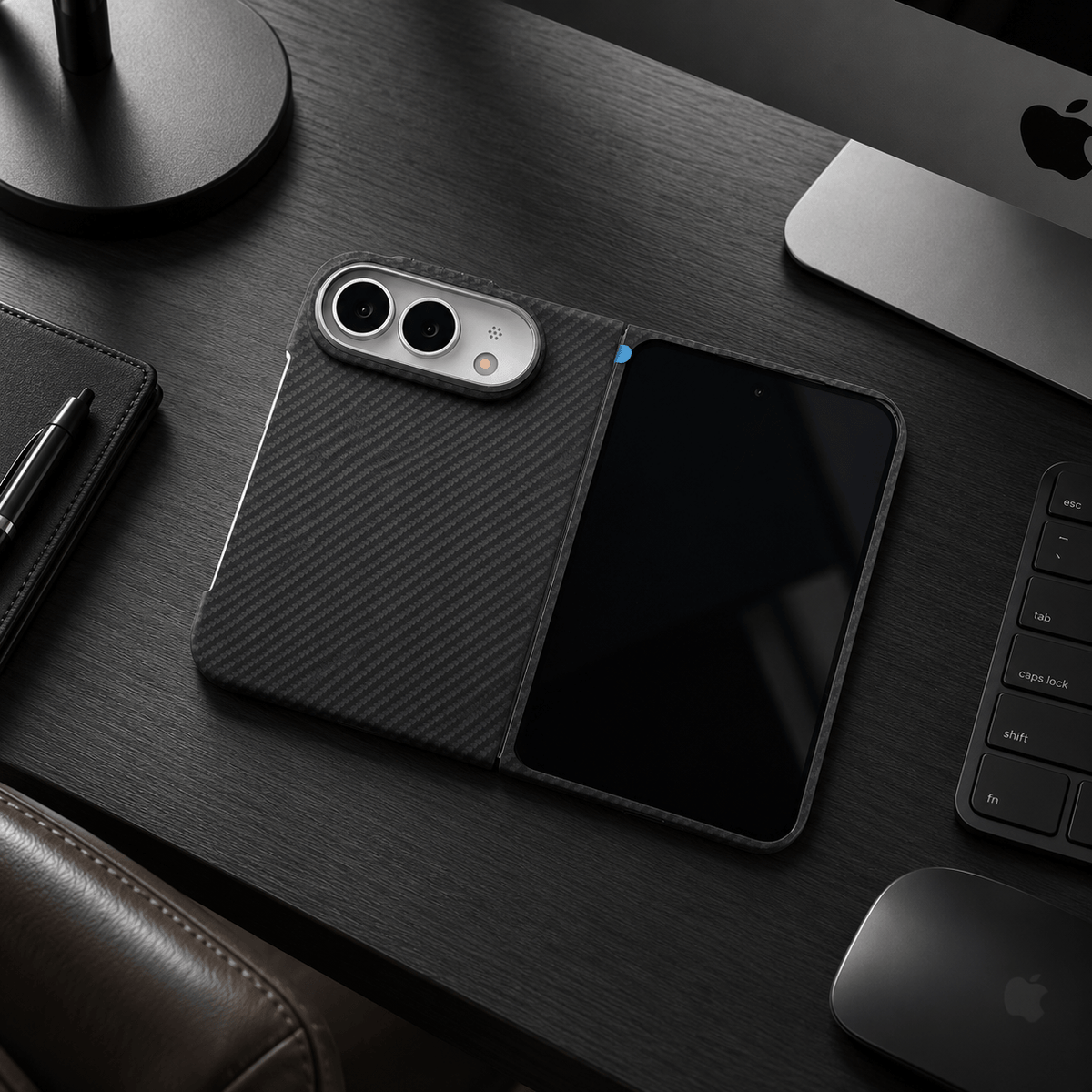 A black carbon-fiber-textured foldable phone case with a built-in screen protector, shown open on a dark wooden desk alongside a laptop, keyboard, and mouse.
