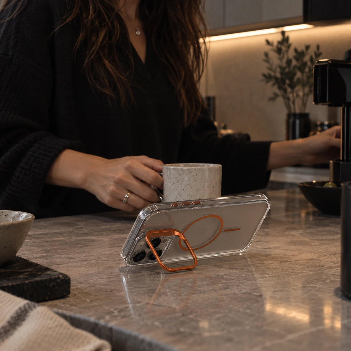 A clear phone case with an orange magnetic ring stand is shown on a kitchen counter, with a person holding a mug in the background.