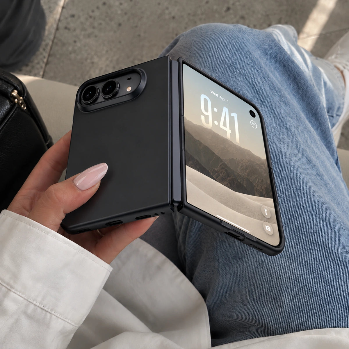 A black protective case for a folding smartphone, shown fitted on a closed Samsung Z Flip phone held in a hand, with the person's blue jeans and white shirt visible in the background.