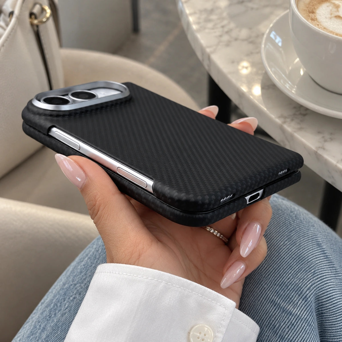 A person holds a black carbon-fiber-textured phone case with a silver metal camera ring in a cafe setting.