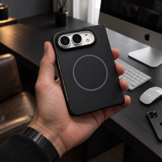 A hand holding a black magnetic phone case with a circular MagSafe ring, shown in a modern office setting with a desk, keyboard, and computer monitor in the background.