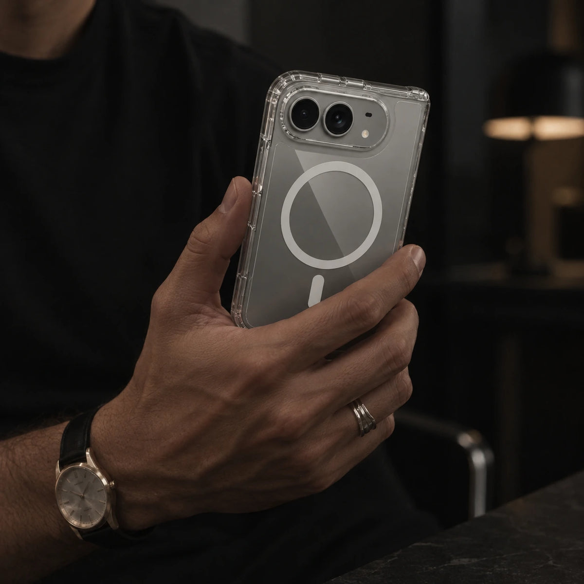 A person holding a smartphone in a dark indoor setting. The phone is fitted with a transparent protective case featuring a prominent white MagSafe-style circular magnetic ring on the back.