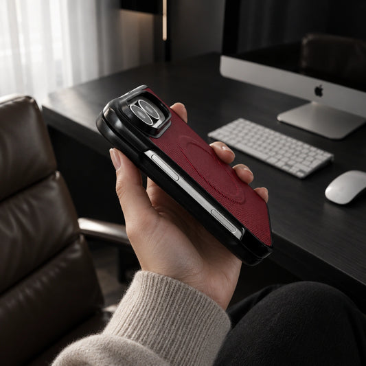 A hand holds a red and black phone case in an office setting with a desk, keyboard, mouse, and computer monitor in the background.